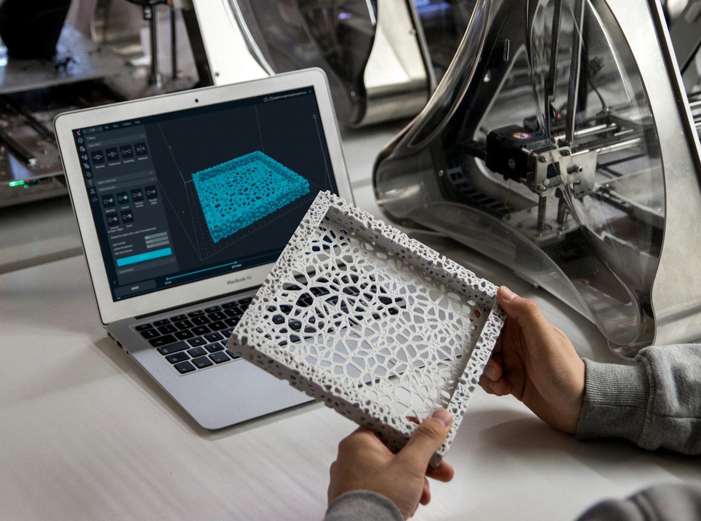 Hände halten ein 3D-gedrucktes Objekt neben einem Laptop, auf dem ein 3D-Modell angezeigt wird.