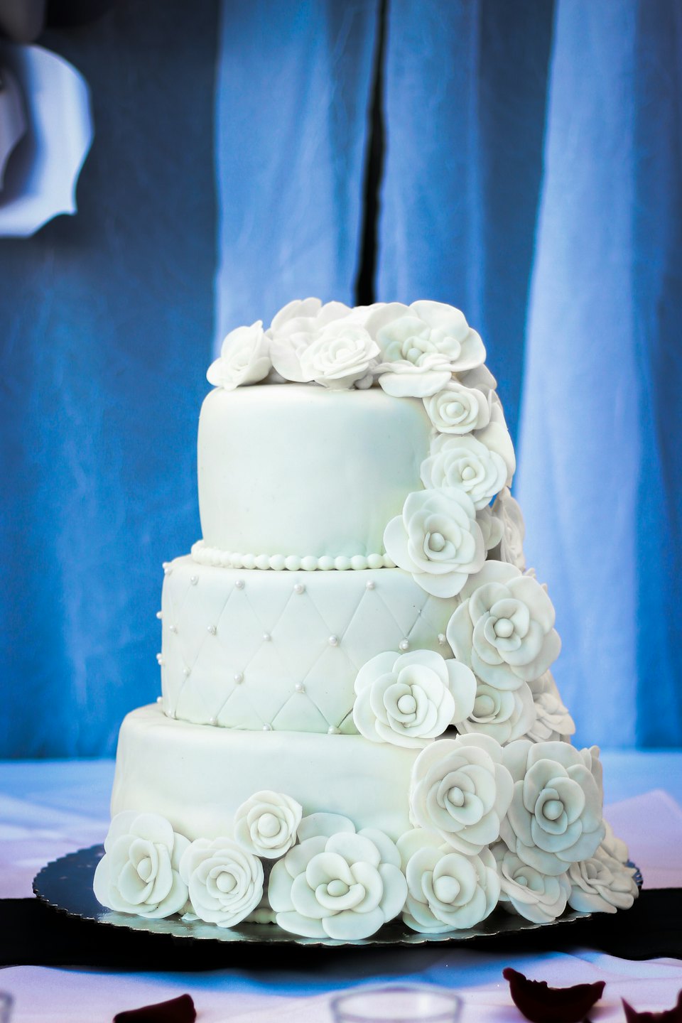 Dreistöckige weiße Hochzeitstorte mit weißen Rosen auf einem silbernen Sockel.